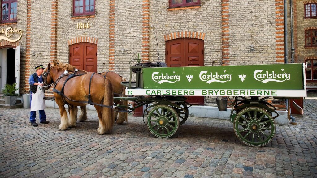 To hester trekker en Carlsberg bryggerivogn foran en historisk murbygning.