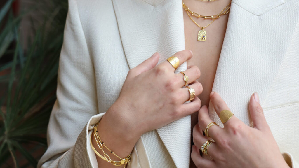 Nahaufnahme einer Person in hellem Blazer mit elegantem Goldschmuck, Ringen, Armband und Halskette.