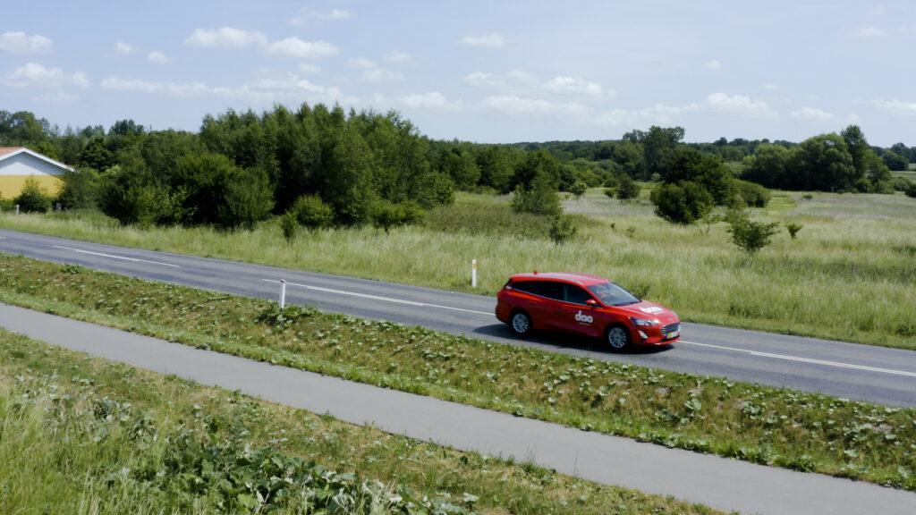 Rotes dao Lieferfahrzeug auf einer Landstraße mit Bäumen und Wiesen im Hintergrund.